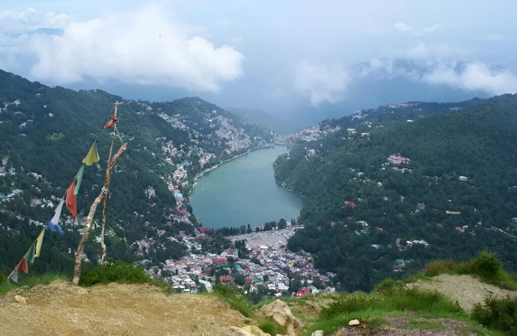 A high-angle view from Naina Peak showing the Naini Lake and the town in a valley surrounded by green hills.