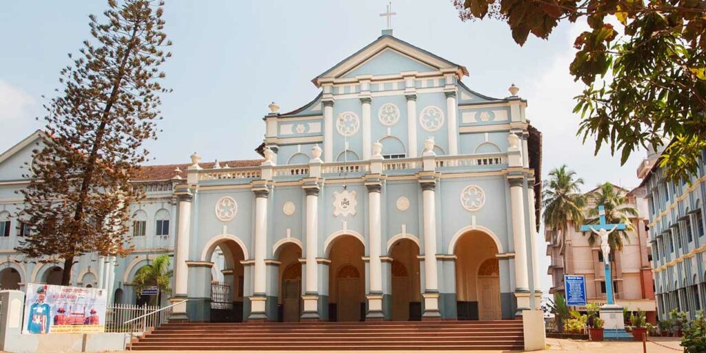 The exterior of St. Aloysius Chapel in Mangalore, with its classical white facade, arched windows, and a statue of St. Aloysius Gonzaga in the landscaped garden at the entrance.